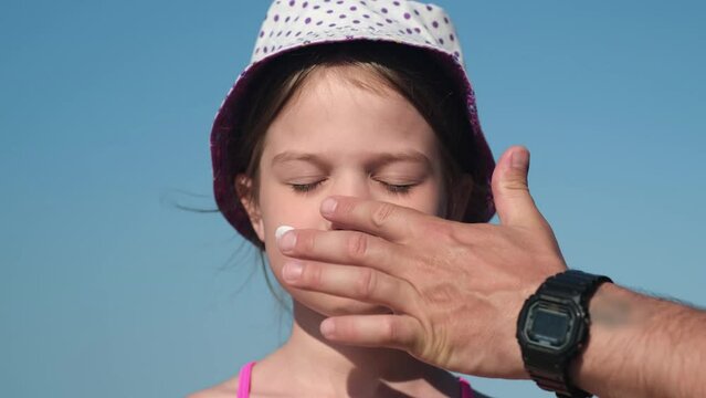 Father applying protective sunscreen on daughter nose at beach on summer holiday vacation. Man hand with care putting sun lotion on child face. Happy little girl with sunblock at sea taking sunbath