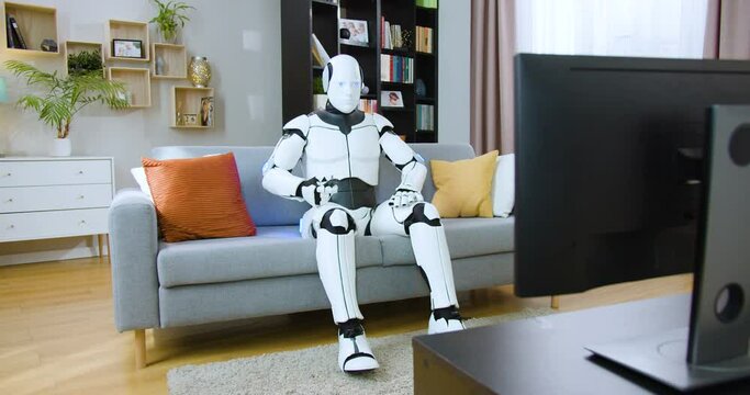Robotic droid relaxing on cozy couch and watching show entertaining programs alone at home. Bionic white cyborg surprising with new information in video on television.