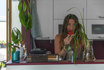 woman eats watermelon in the kitchen behind the counter. In front of the woman on the counter are flowers and coffee beans in a glass jar. Behind the woman are white kitchen cabinets.
