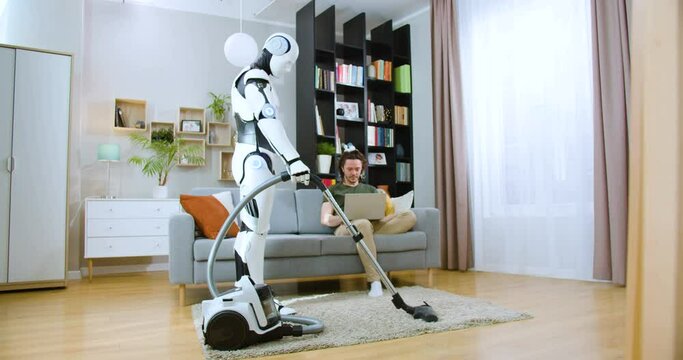 Young caucasian man sitting on comfy sofa while computerized cyborg vacuuming floor in apartment with modern design. Handsome male freelancer working on wireless laptop at home.