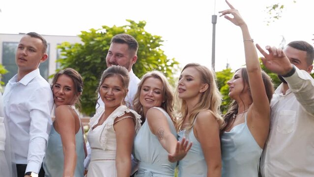 Groom, bride and excited friends cheering on camera on wedding day in park. Groomsmen and pretty bridesmaids in identical dresses have fun waving hand and posing, dancing. Slow motion.