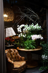 Florist shop window. Beautiful decoration of luxurious white flowers, interior items, lighting. Showcase window with atmospheric lights, vintage armchair. Dark exquisite stage photo. Vertical image 
