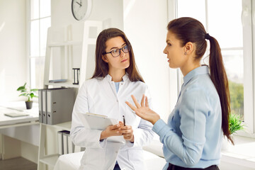 Fototapeta premium Young Caucasian woman in nurse coat works as doctor in cosmetology clinic listens to female patient who has applied for consultation in field of beauty stand in spacious bright office of hospital