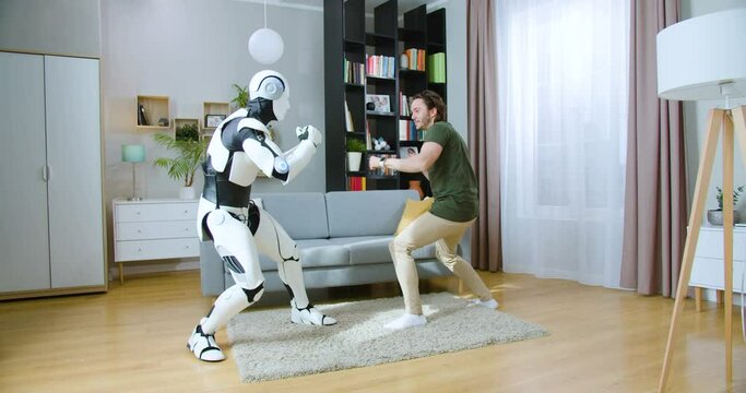 Muscular caucasian man boxing with humanoid cyborg while practicing punches in living room. Futuristic bionic robot having battle with young man at home.