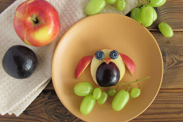 Funny owl made from fruits. Next to the plate are fruits: an apple, a plum and a grape.
