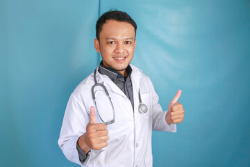 Portrait of a young Asian man doctor, a medical professional is smiling and showing thumbs up or OK sign isolated over blue background