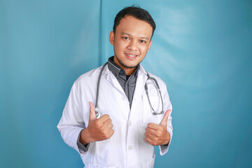 Portrait of a young Asian man doctor, a medical professional is smiling and showing thumbs up or OK sign isolated over blue background