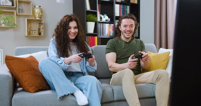 Happy girlfriend and boyfriend playing online on television console while sitting on comfortable sofa. Cheerful caucasian couple using joysticks for gaming challenge competition.