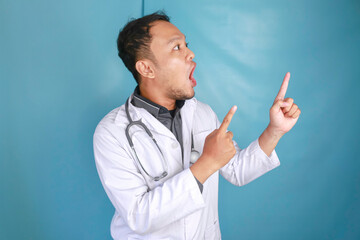 Young Asian man doctor, a medical professional is shock and pointing upward at a copy space isolated over blue background