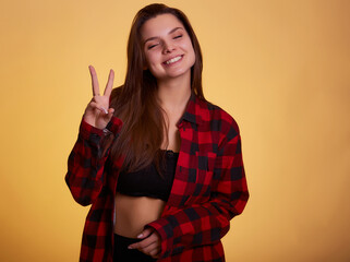 Beautiful lady hold arm hand fingers show v-sign say hi friends relatives positive mood wear casual checkered plaid shirt clothes outfit isolated yellow background.