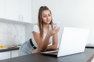 Young woman uses smartphone in the kitchen with laptop on the countertop.