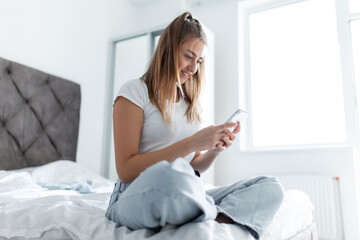Young hipster woman using smartphone while sitting on bed in bright room