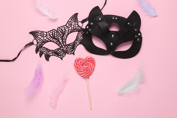 Lace and leather cat masks with lollipop and feathers on a pink background. Top view. flat lay