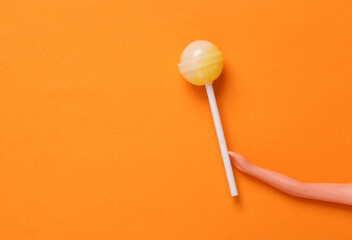 Doll hand holding lollipop on orange background. Minimal halloween, junk food, sweets, concept. Top view