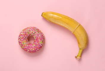 Sex education, food concept. Ripe banana with condom and donut on pink background. Top view