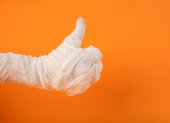 Mummy's hand wrapped in white bandages shows a thumbs up on an orange background. Happy Halloween....