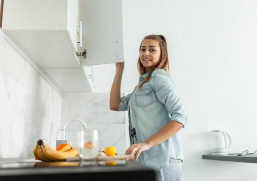 Cute Young Woman Opens The Kitchen Cabinet Door. Healthy Food Concept