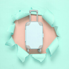 Toy luggage through a torn hole on blue-pink pastel background. Travel concept. Pastel color trend....