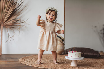 Cute one year girl in beige dress and cute flower crown posing with white birthday cake in the studio. Party concept