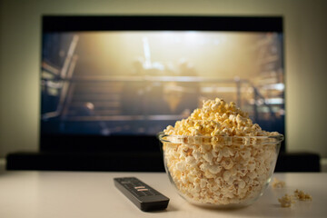 A glass bowl of popcorn and remote control in the background the TV works. Evening cozy watching a...