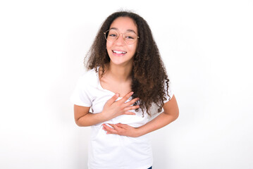 young beautiful girl with afro hairstyle wearing white t-shirt over white wall keeps hands crossed, laughs out loud at good joke, wears casual clothes.