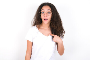 young beautiful girl with afro hairstyle wearing white t-shirt over white wall being in stupor shocked, has astonished expression pointing at oneself with finger saying: Who me?