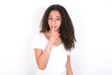 young beautiful girl with afro hairstyle wearing white t-shirt over white wall makes hush gesture, asks be quiet. Don't tell my secret or not speak too loud, please!