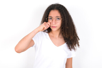 Unhappy young beautiful girl with afro hairstyle wearing white t-shirt over white wall crying while...