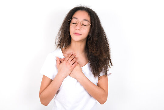 young beautiful girl with afro hairstyle closes eyes and keeps hands on chest near heart, expresses sincere emotions, being kind hearted and honest. Body language and real feelings concept.