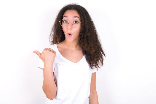 Shocked young beautiful girl with afro hairstyle wearing white t-shirt over white wall points with thumb away, indicates something. Check this out. Advertisement concept.