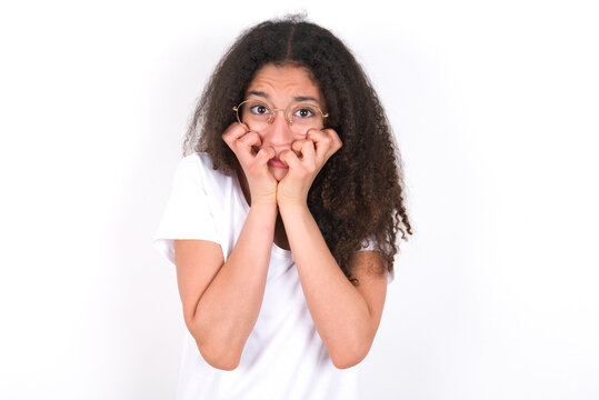 Anxiety - young beautiful girl with afro hairstyle wearing white t-shirt over white wall covering his mouth with hands scared from something or someone bitting nails.