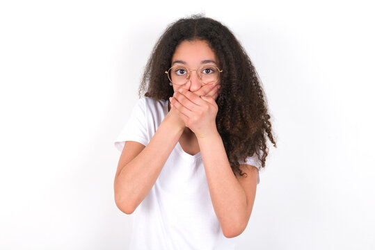 Upset young beautiful girl with afro hairstyle wearing white t-shirt over white wall, covering her mouth with both palms to prevent screaming sound, after seeing or hearing something bad.