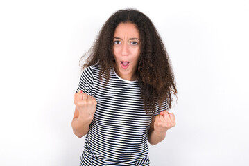 Joyful excited lucky young beautiful girl with afro hairstyle wearing striped t-shirt over white wall cheering, celebrating success, screaming yes with clenched fists