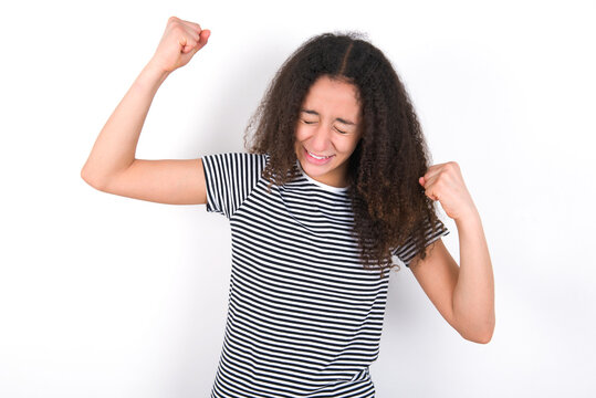 Attractive young beautiful girl with afro hairstyle wearing striped t-shirt over white wall celebrating a victory punching the air with his fists and a beaming toothy smile.