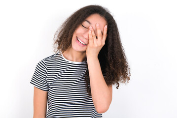young beautiful girl with afro hairstyle wearing striped t-shirt over white wall makes face palm and smiles broadly, giggles positively hears funny joke poses