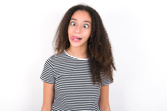 Funny young beautiful girl with afro hairstyle wearing striped t-shirt over white wall makes grimace and crosses eyes plays fool has fun alone sticks out tongue.