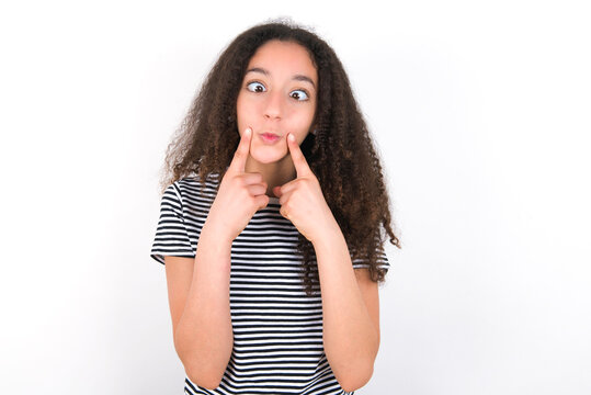 young beautiful girl with afro hairstyle wearing striped t-shirt over white wall crosses eyes and makes fish lips funny grimace