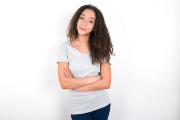 Confident young beautiful girl with afro hairstyle wearing grey t-shirt over white wall with arms crossed looking to the camera