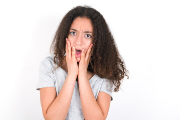 Upset young beautiful girl with afro hairstyle wearing grey t-shirt over white wall touching face with two hands
