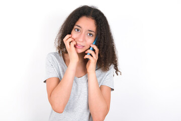 Afraid funny young beautiful girl with afro hairstyle wearing grey t-shirt over white wall holding telephone and bitting nails
