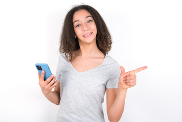 Astonished young beautiful girl with afro hairstyle wearing grey t-shirt over white wall holding her telephone and pointing with finger aside at empty copy space