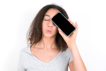 Fototapeta premium Adorable young beautiful girl with afro hairstyle wearing grey t-shirt over white wall holding modern device covering eye with lips pouted