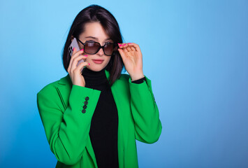 Charming brunette woman uses mobile phone, looking at camera wears in green blazer and sun glasses isolated over blue background. Be communicated