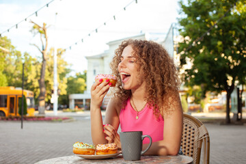 young attractive curly woman sits at table in cafe on summer terrace. Colorful delicious donuts,...