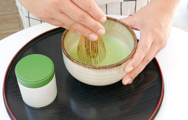 Hand hold whisk preparing and mixing traditional organic Japanese matcha green tea powder with milk (cold whisk) in a bowl on black tray