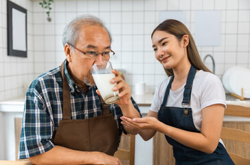 Asian lovely family, young daughter prepare breakfast for older father. Attractive female wear apron bake bread serve with milk to senior elderly dad sitting on eating table in kitchen at house.