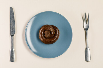 Plastic excrement lies on a plate between a fork and a knife close-up. The concept of an inept cook, fake news, shitty restaurant, bad food, taste of shit