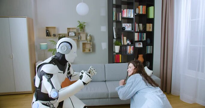 Caucasian woman imitating punches with humanoid cyborg while practicing boxing in living room. Futuristic bionic robot having battle with young lady at home.