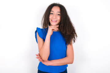 Optimistic Teenager girl with afro hairstyle wearing blue T-shirt over white wall  keeps hands partly crossed and hand under chin, looks at camera with pleasure. Happy emotions concept.