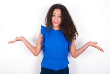 Clueless Teenager girl with afro hairstyle wearing blue T-shirt over white wall  shrugs shoulders with hesitation, faces doubtful situation, spreads palms, Hard decision